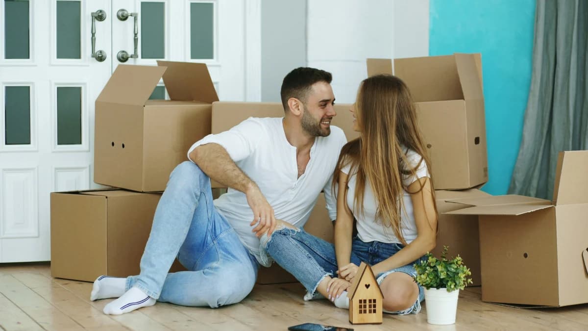 Un couple assis au sol dans une pièce entourée de cartons de déménagement.